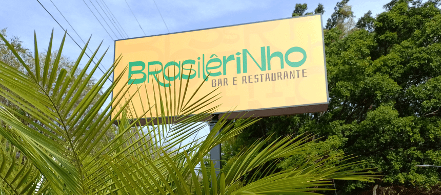 Foto do luminoso da entrada do Brasilêrinho Bar e Restaurante. Na frente do luminoso há algumas folhas de coqueiro e ao fundo, várias árvores.